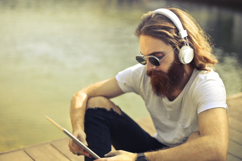 A man with long brown hair sits on a dock listening to an audiobook